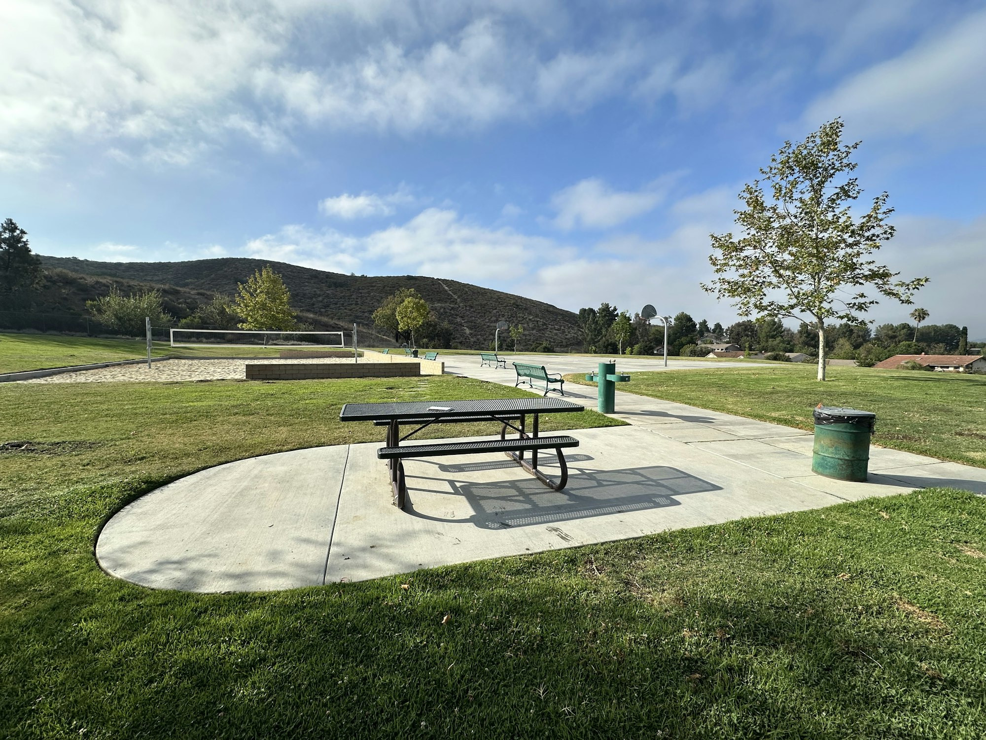A peaceful park scene with picnic tables, a volleyball court, grassy areas, and trees under a cloudy sky.