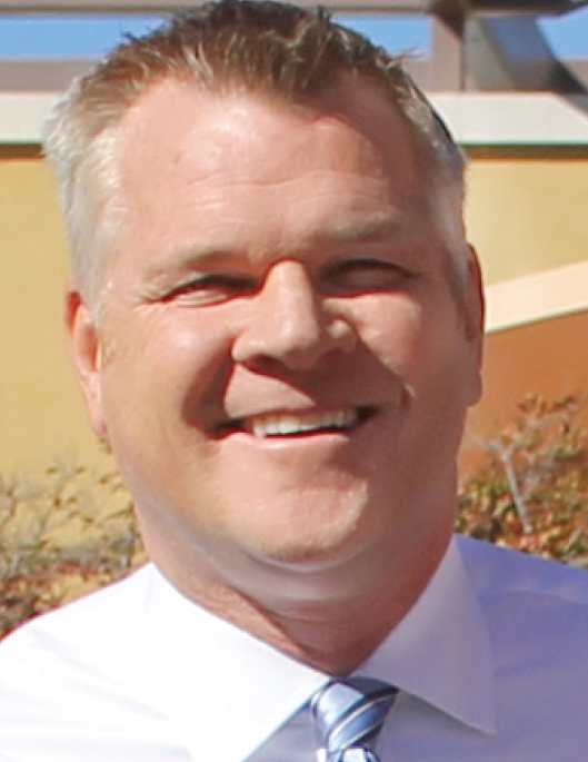 A person with a short haircut, smiling, wearing a white shirt and striped tie, standing outdoors.