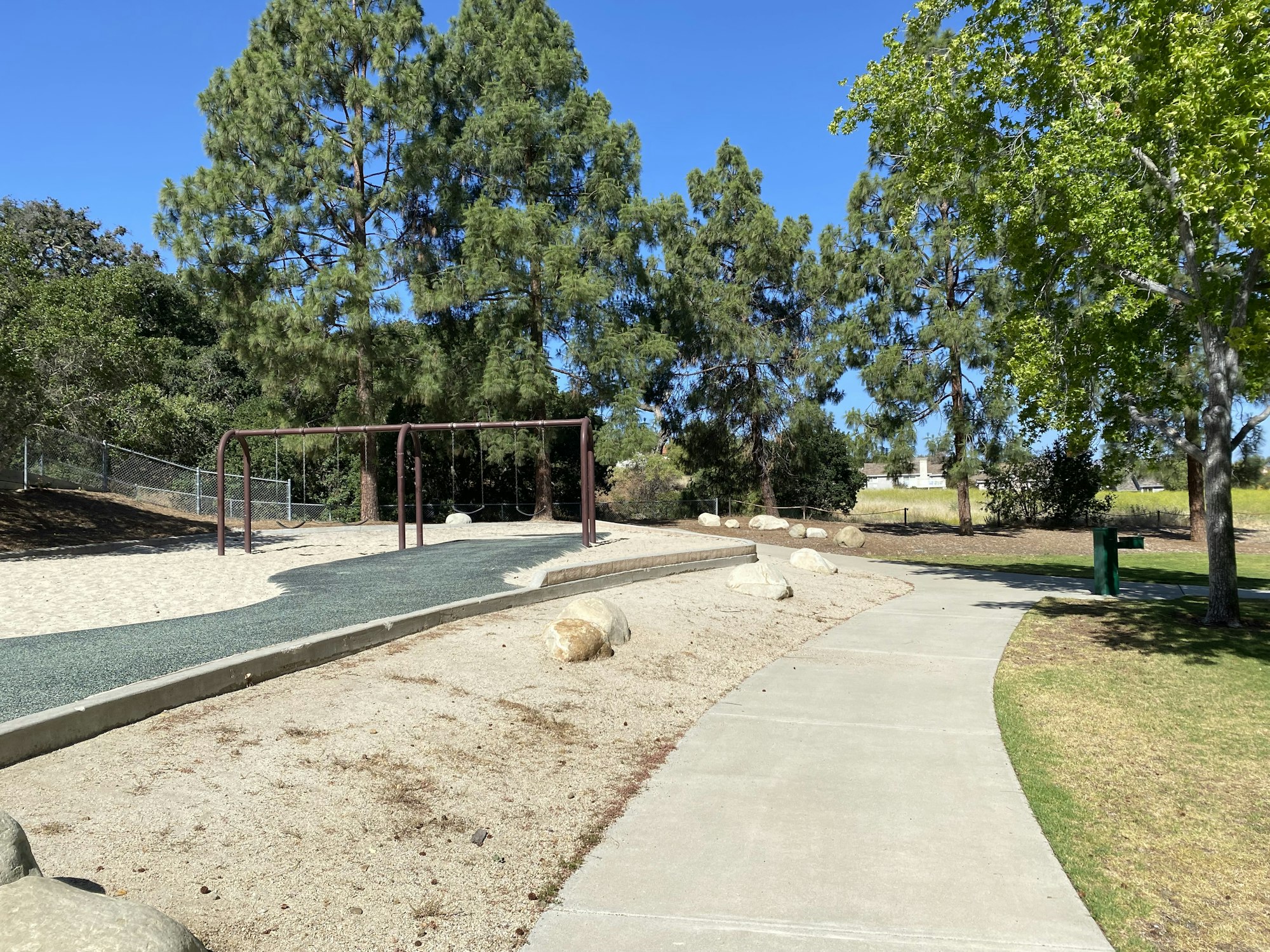 Playground swing set, sand area, walking path, and trees on a sunny day.
