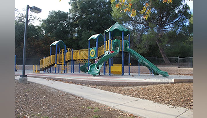 A playground with yellow and green structures, slides, and stairs, surrounded by trees and a fence.
