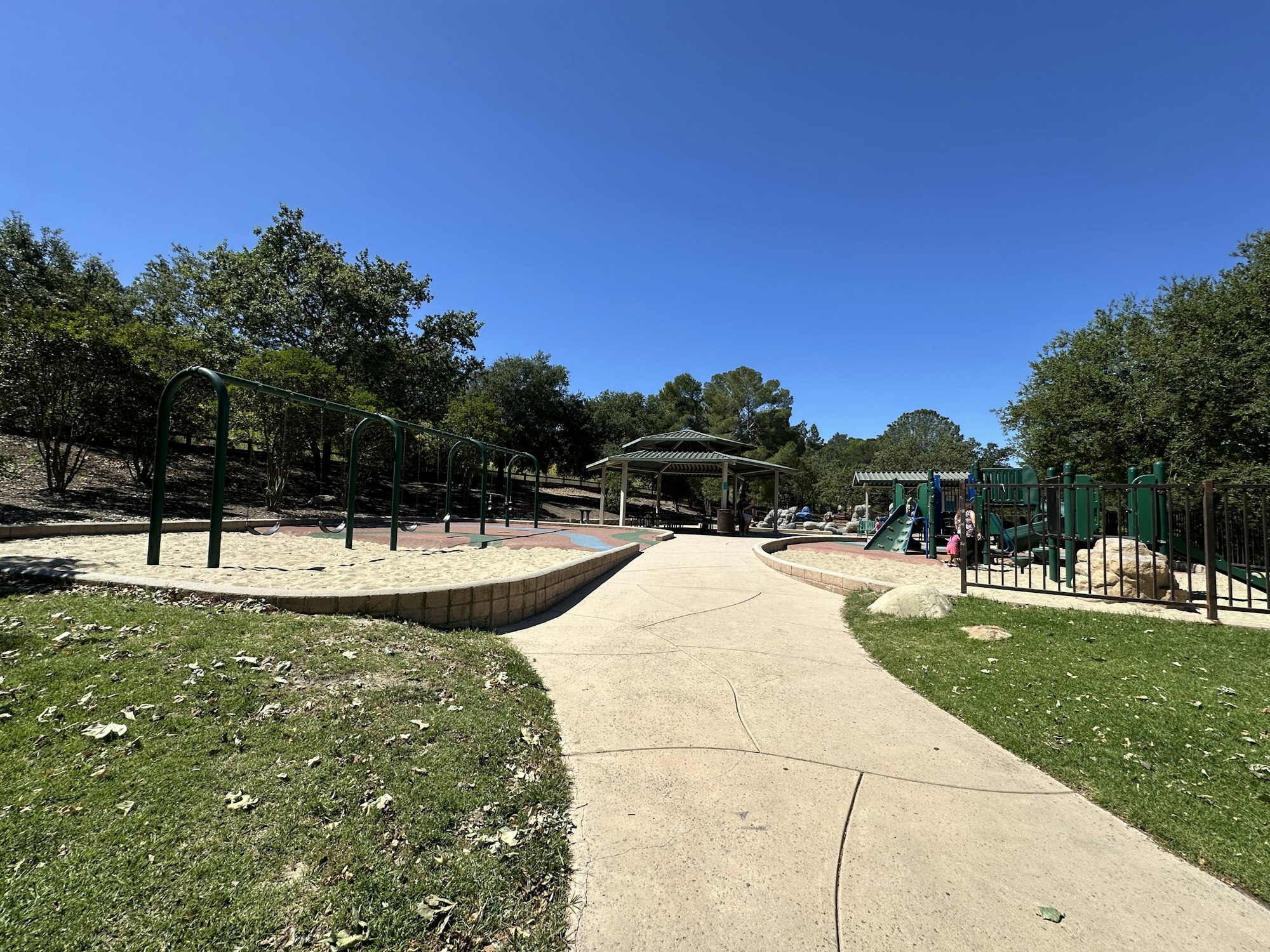 A sunny playground featuring swings, play structures, and a shaded area, surrounded by trees and a clear blue sky.