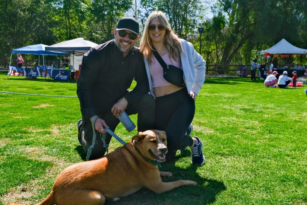 Two people kneeling and smiling with a dog on grass at an outdoor event with tents in the background.