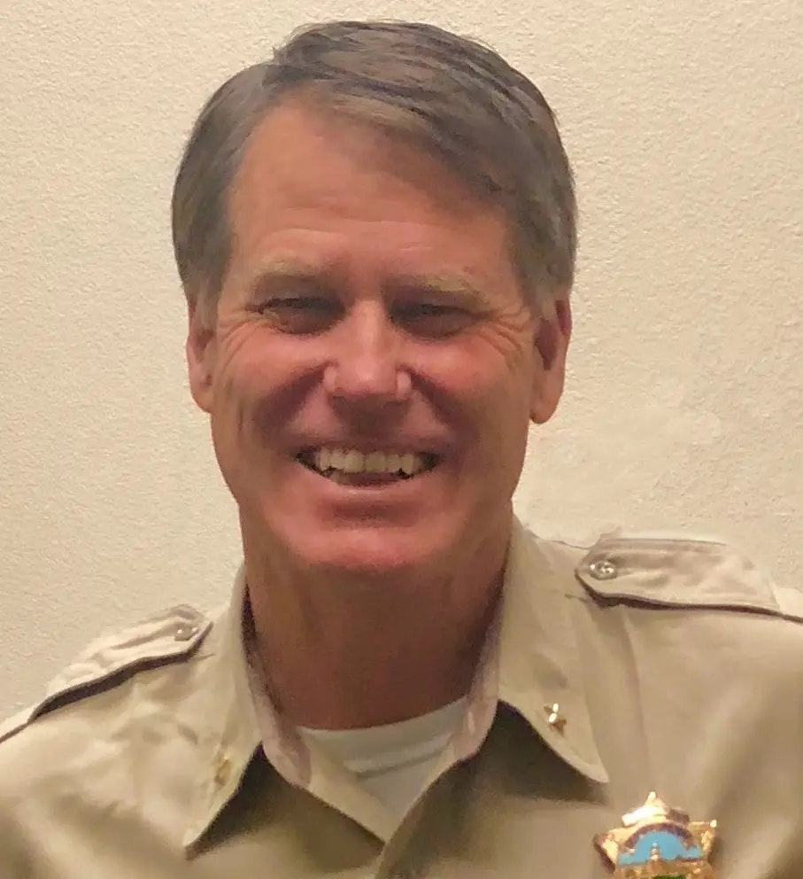 A person in a uniform with a badge, smiling, against a plain background.