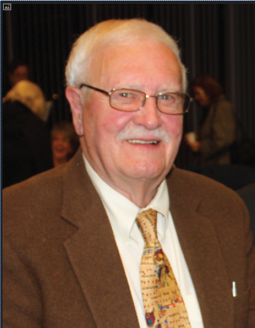 An elderly person wearing glasses and a brown suit with a tie, smiling at an event.
