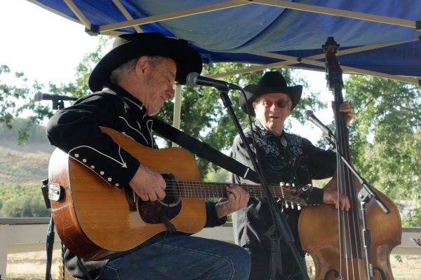 Two musicians in cowboy hats, one playing a guitar and singing, the other on a double bass, performing outdoors under a blue canopy.