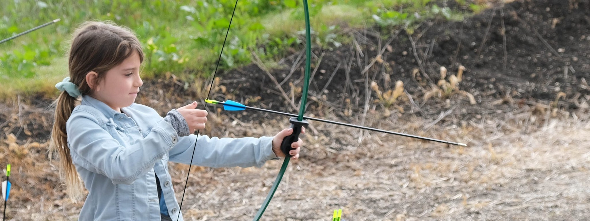 A young girl practicing archery, aiming an arrow with a bow outdoors.