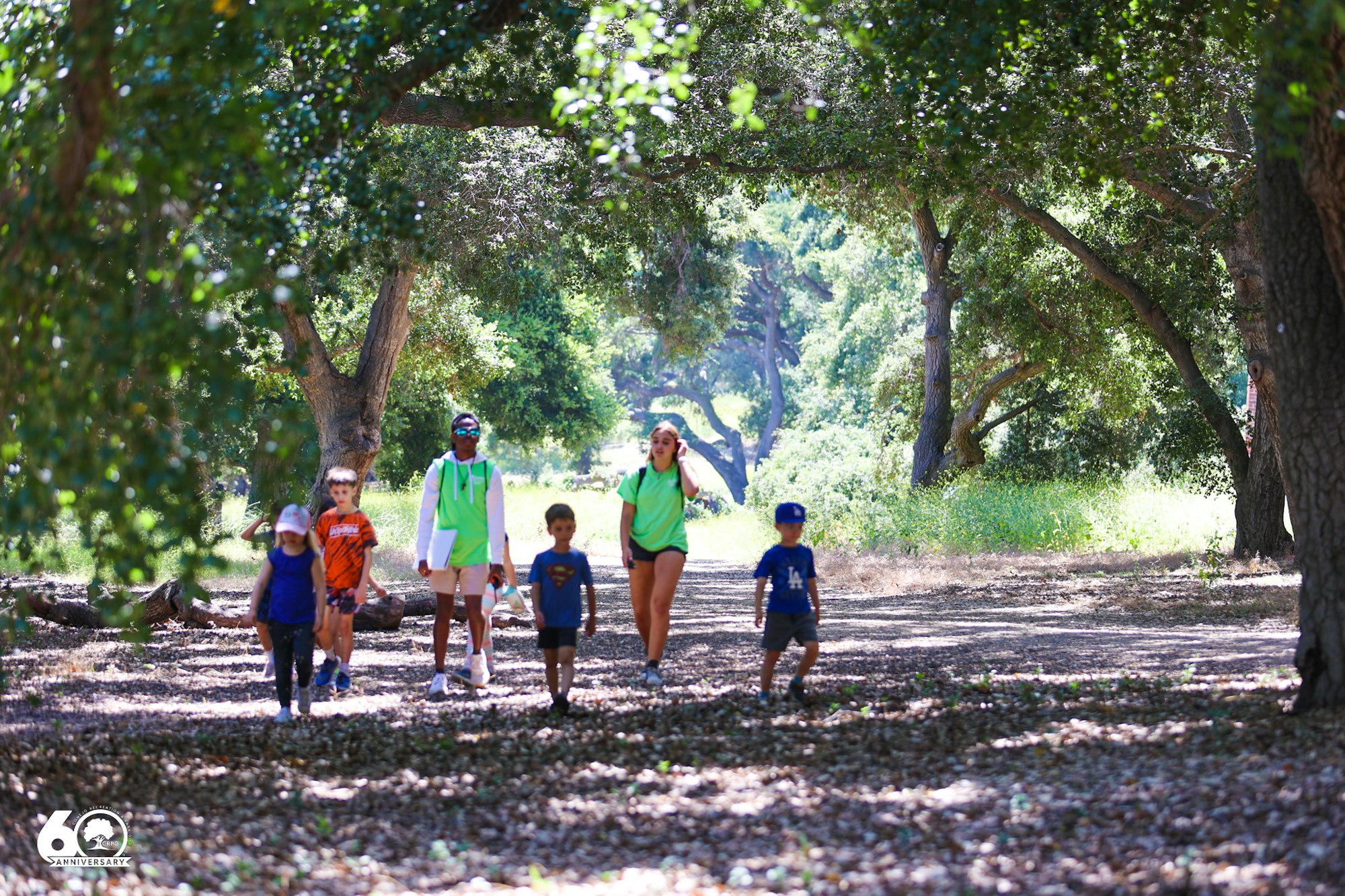 A group of people, including children and adults, walking on a leaf-covered path in a forested area, surrounded by trees and greenery.