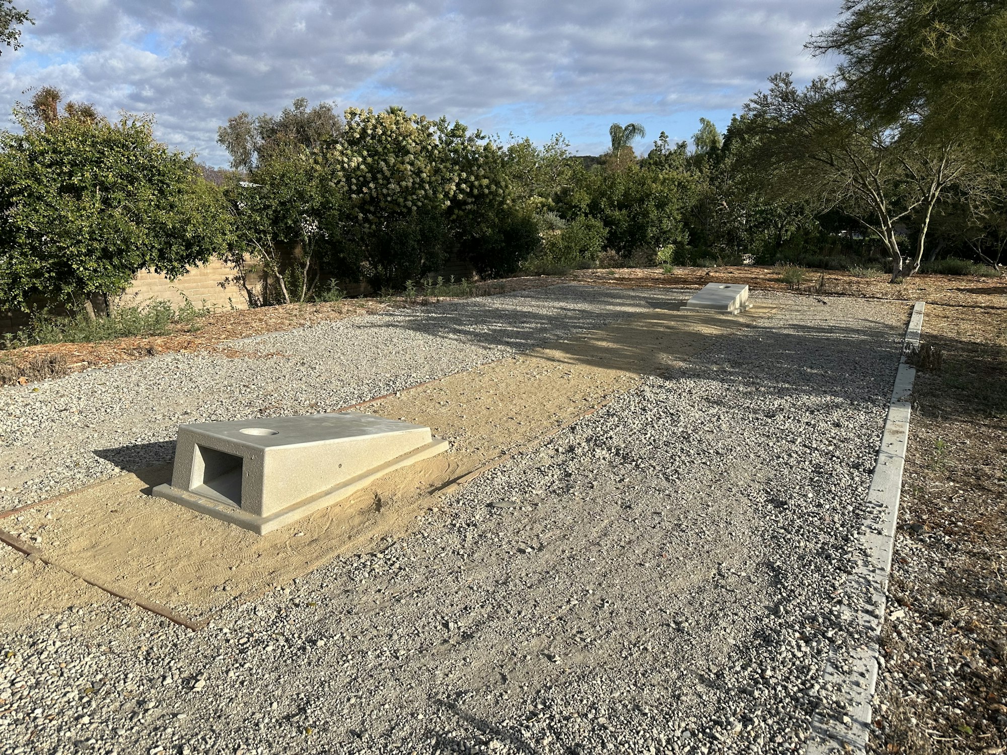 The image shows a gravel area with two concrete structures and surrounding greenery under a cloudy sky.