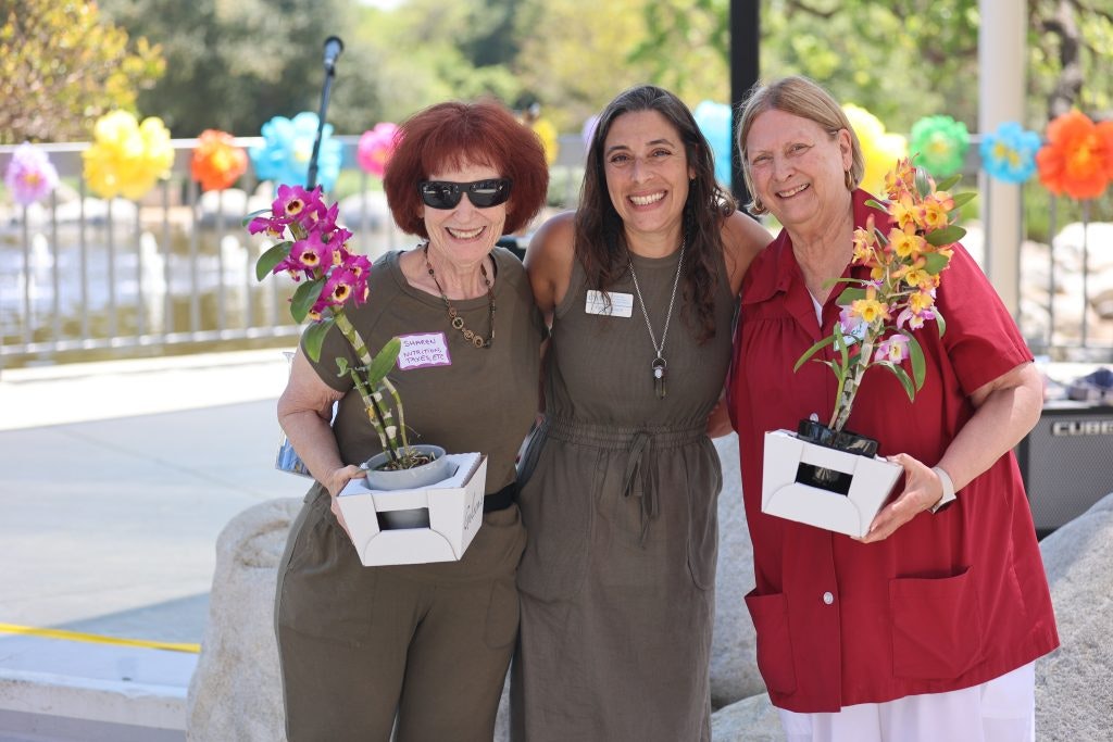 Three people smiling, standing outdoors, holding flowers in pots with colorful decorations in the background.