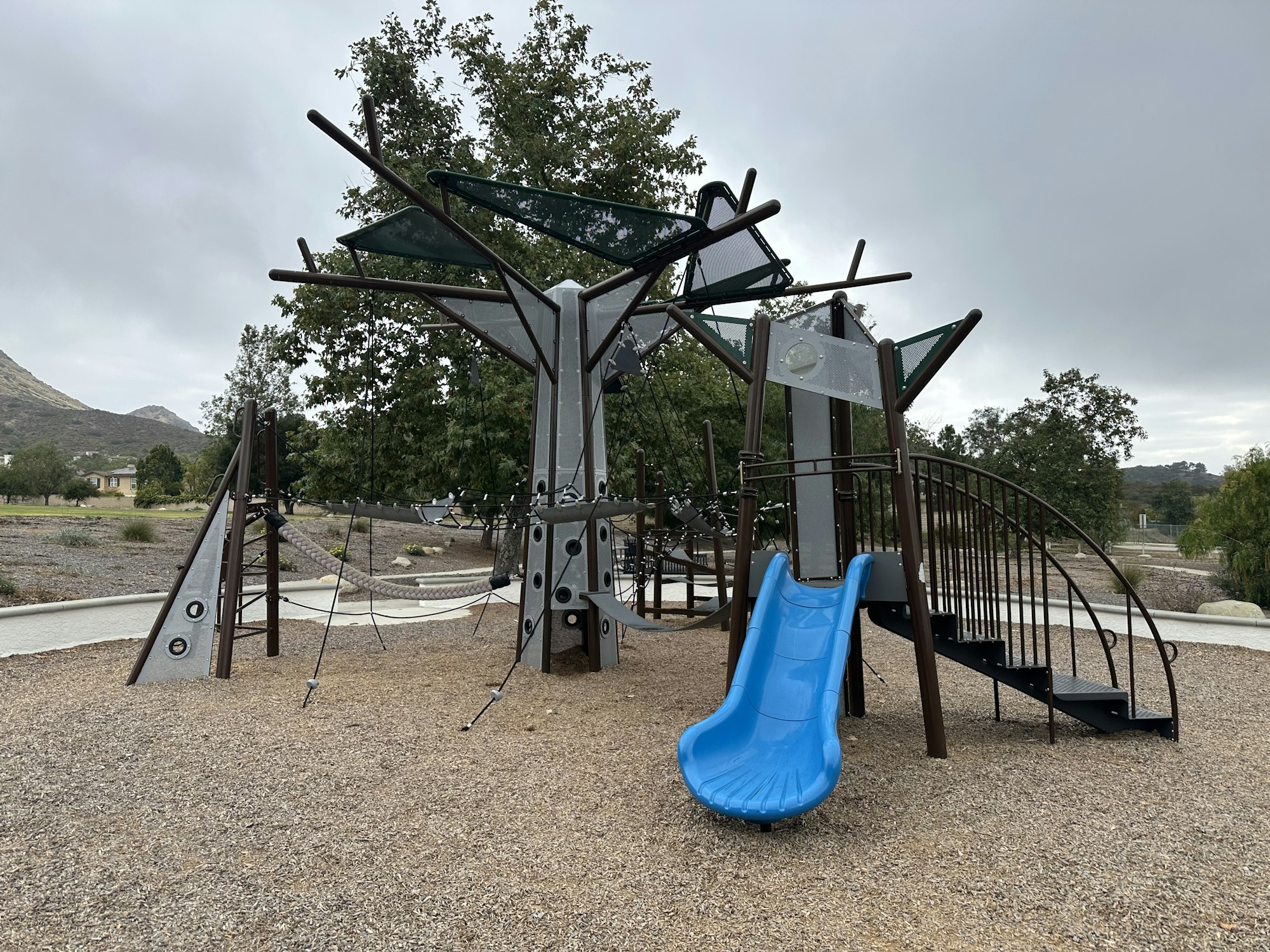 A modern playground with a blue slide, climbing structures, and a spiral staircase, set in a park environment.