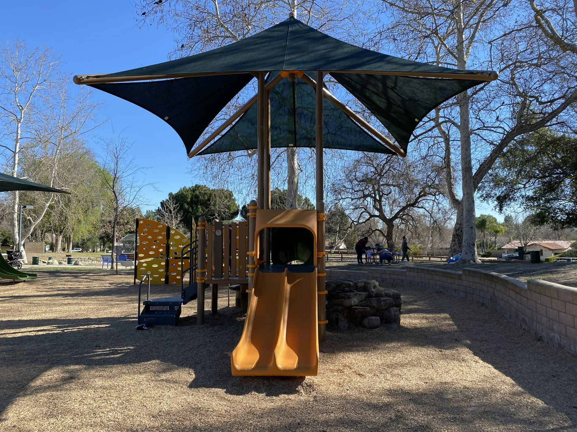 Playground with slides, climbing wall, and shade canopy; trees and people in the background.