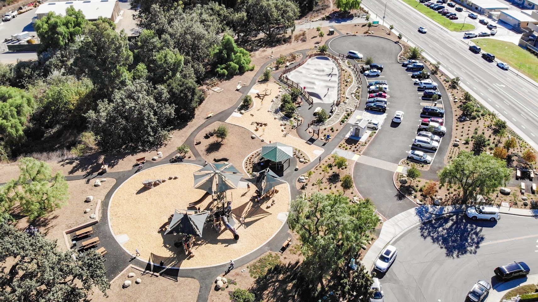 Aerial view of a park with playground structures, pathways, trees, and adjacent parking lot.