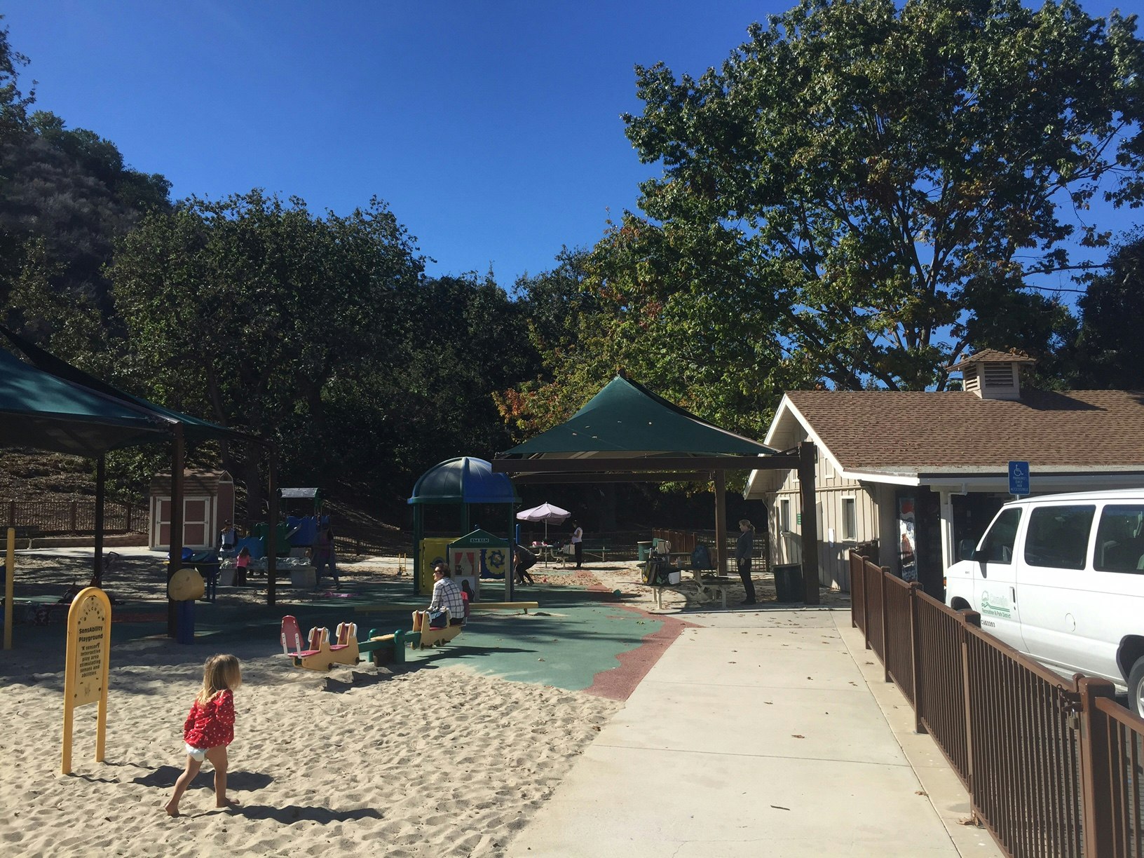 Children playing in a sunny outdoor playground with a sandbox, seesaw, and nearby building under trees.