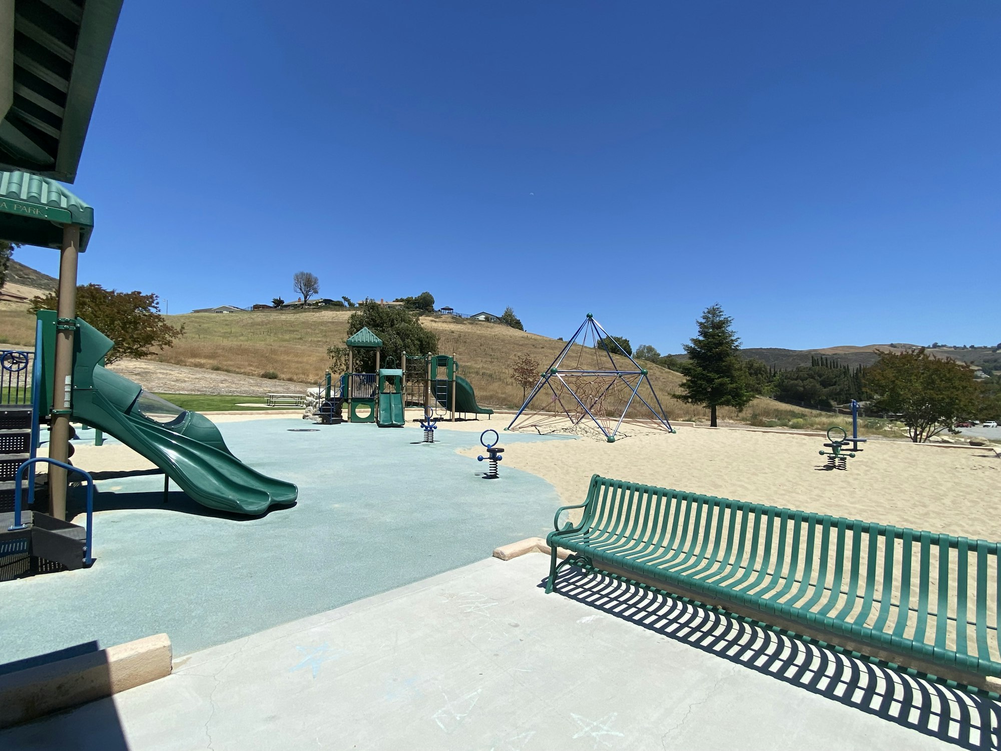 Playground with green slides, climbing structure, benches, and hills in the background under a clear blue sky.