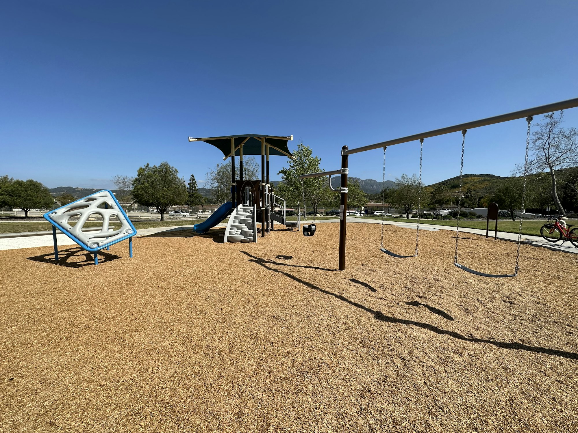 Playground with swings, climbing equipment, slide, and a canopy, surrounded by trees and open space on a sunny day.