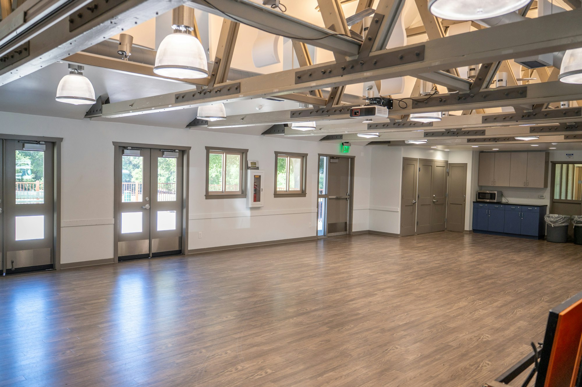 Spacious room with wooden floor, ceiling lights, windows, doors, and a small kitchen area with cabinets and a microwave.