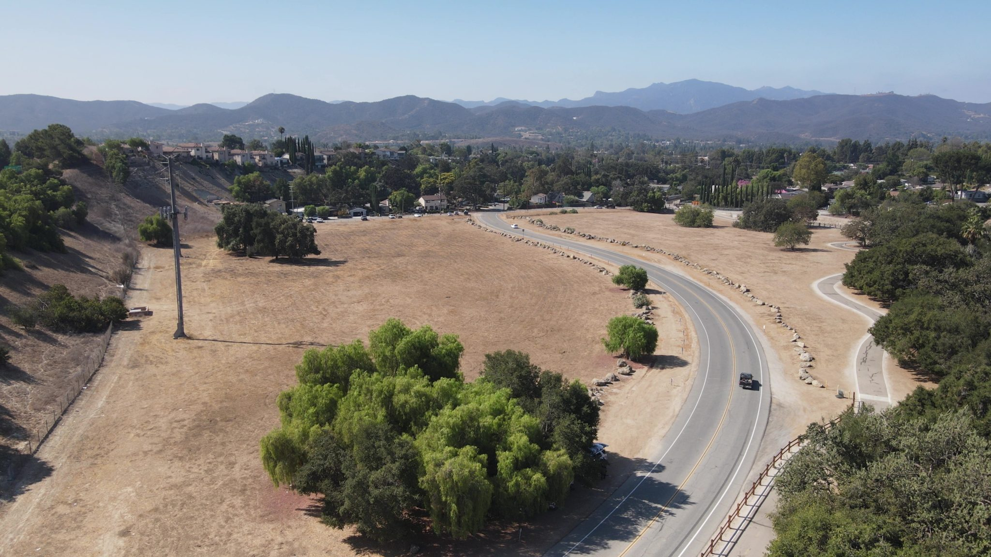 Winding road through a dry landscape, with scattered trees, hills, and distant mountains under clear skies.