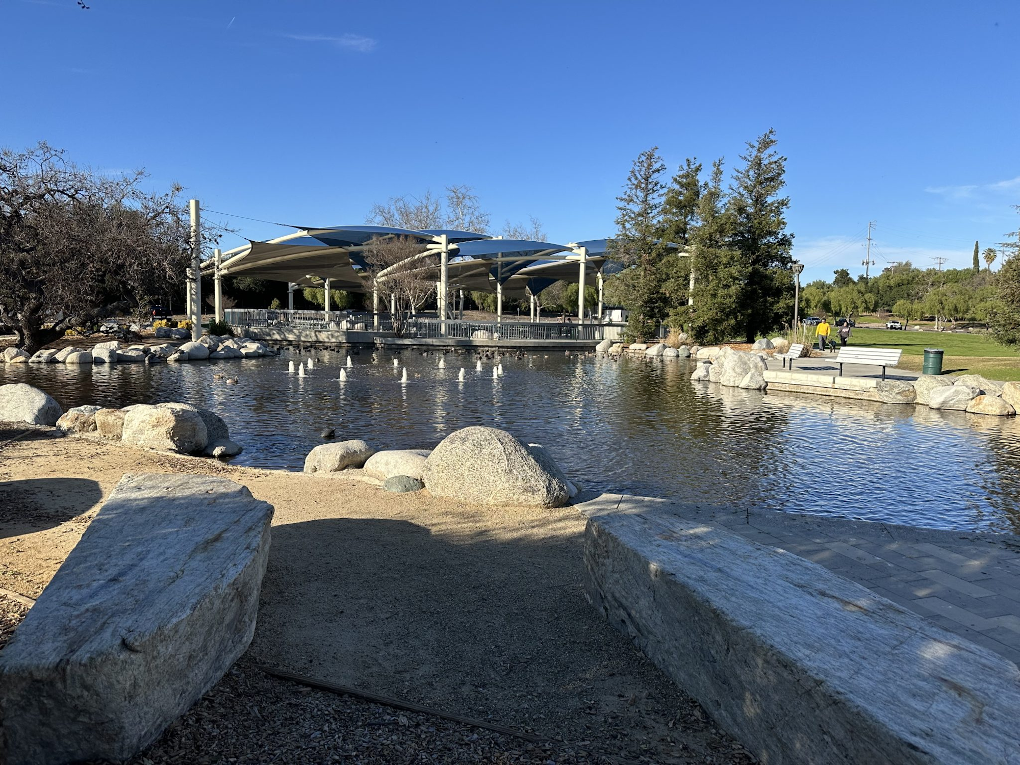 Park with a pond, rocks, trees, and a shaded pavilion under a clear blue sky.