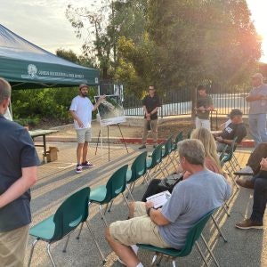 An outdoor meeting with a group of people, a speaker presenting under a canopy, and an audience seated in chairs.