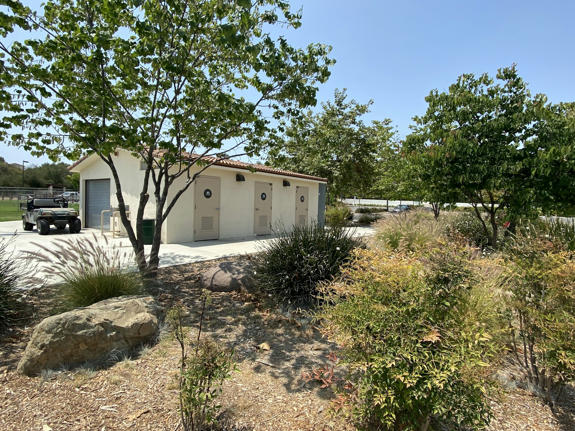 Restroom building in a park setting with trees, bushes, and a utility vehicle nearby.