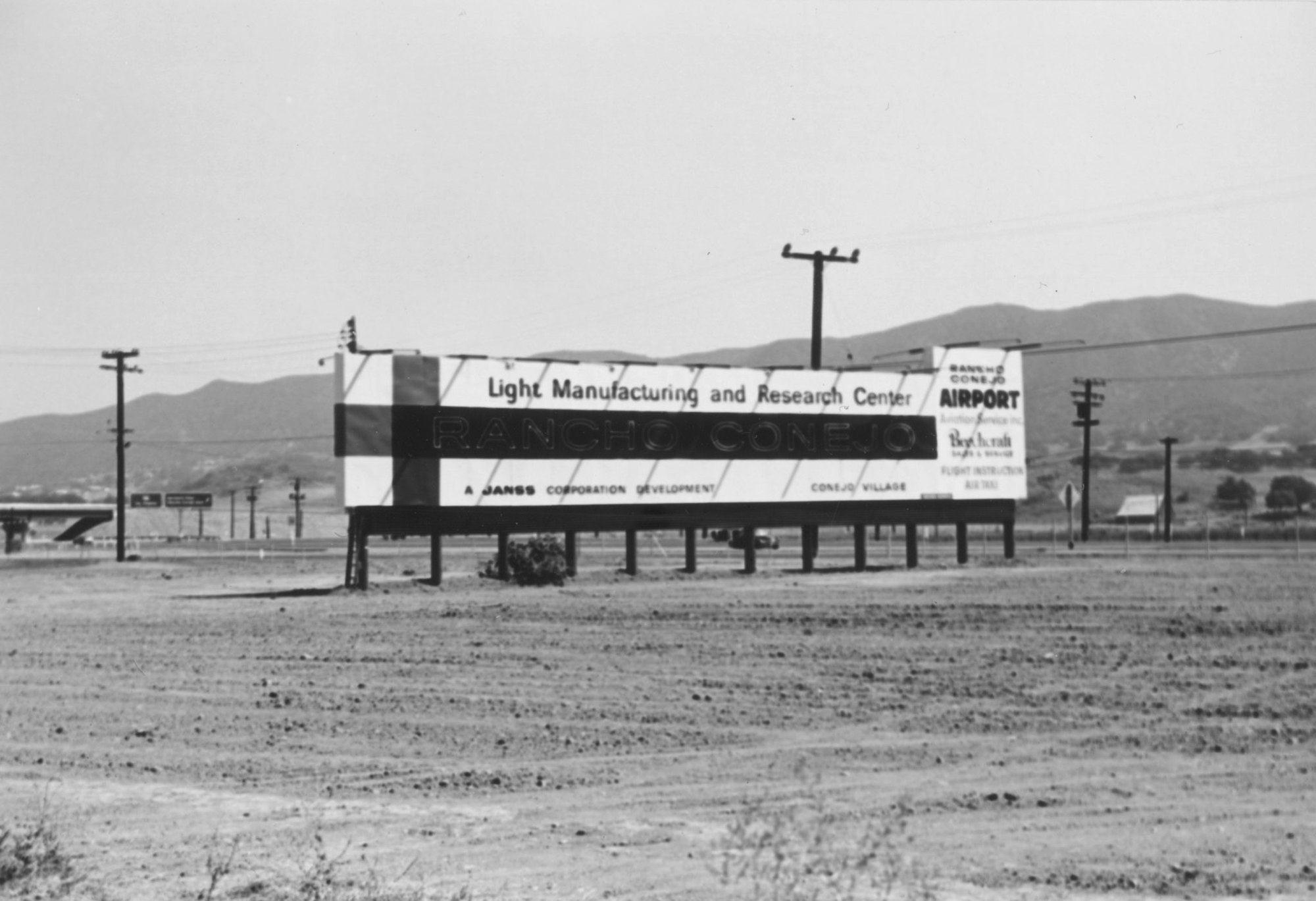A billboard promoting Rancho Conejo Light Manufacturing and Research Center, and airport, in a barren landscape with mountains.