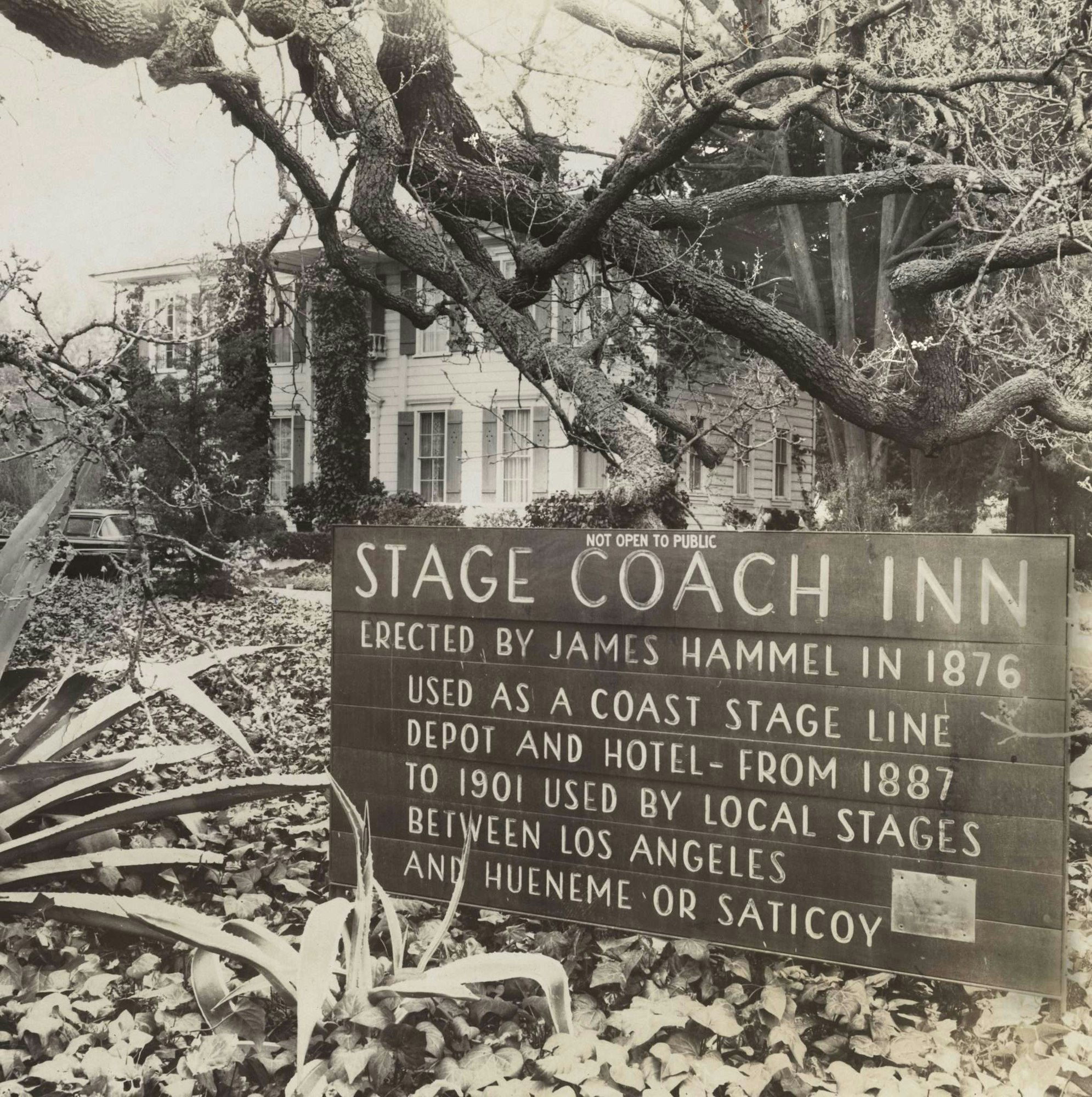 Stage Coach Inn sign with historical info, surrounded by trees and plants, in front of a two-story building.