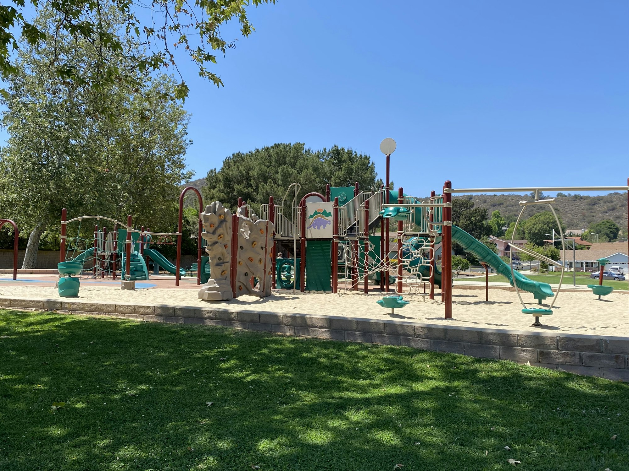 Playground with slides, climbing structures, and sand, surrounded by trees and grass under a clear blue sky.
