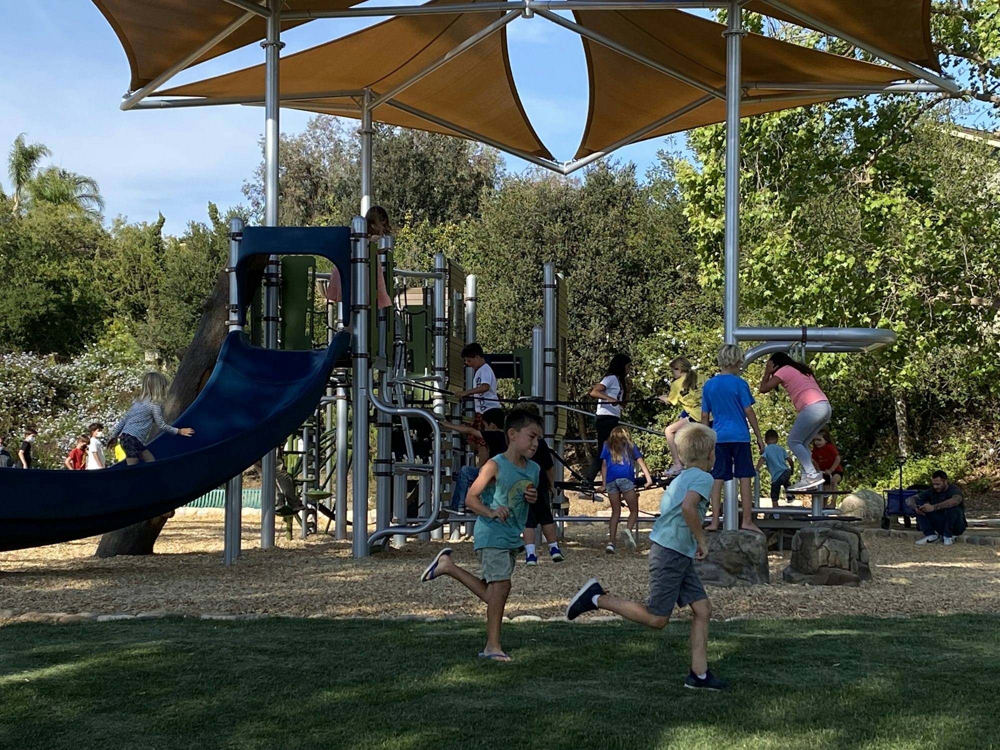 Children playing on a playground with slides and climbing equipment, surrounded by trees and shade structures.