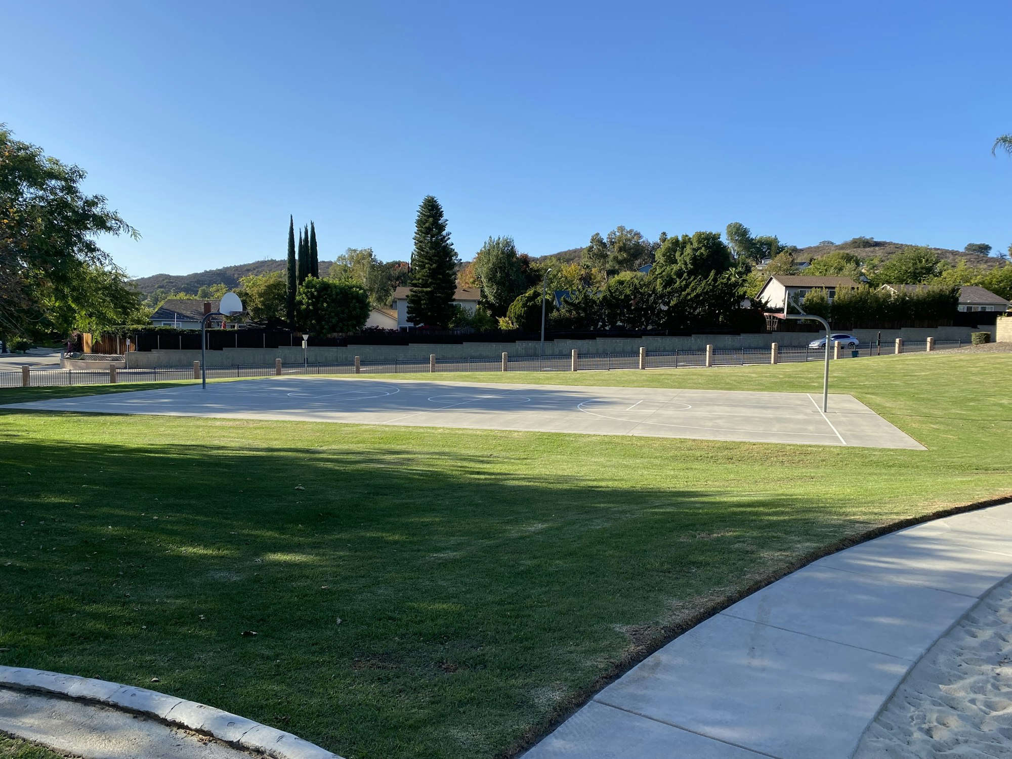 Outdoor basketball court surrounded by grass, trees, and a sidewalk under a clear blue sky.