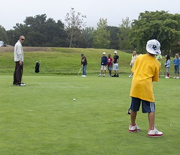 A group of people, including a child in yellow, are on a golf course practicing putting.