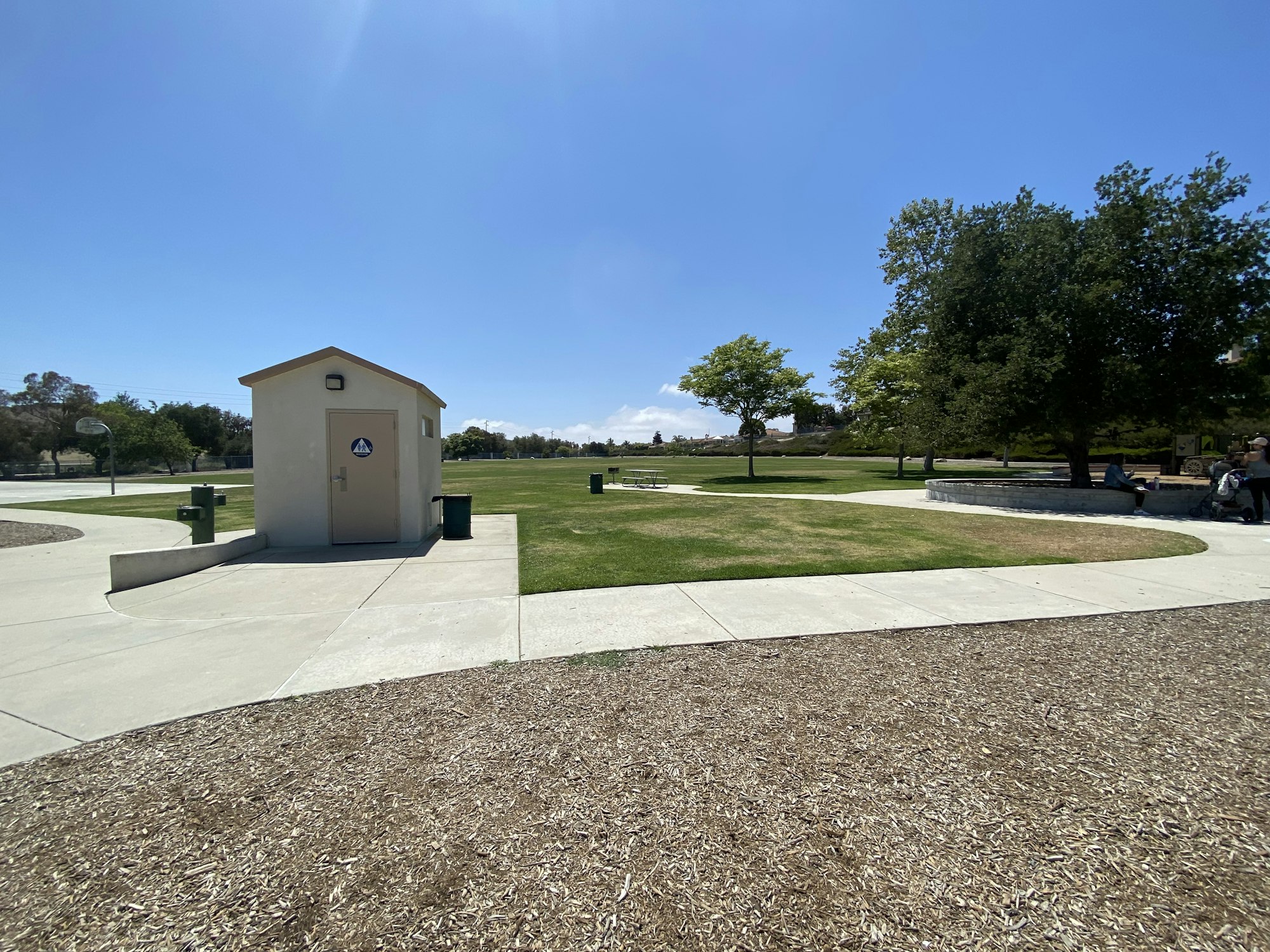 A park with a restroom building, pathways, trees, and people sitting on a bench under a tree.