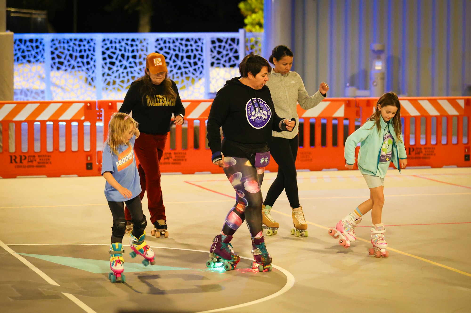 A group of people, including children and adults, is roller skating on a rink, surrounded by safety barriers and colorful lights.
