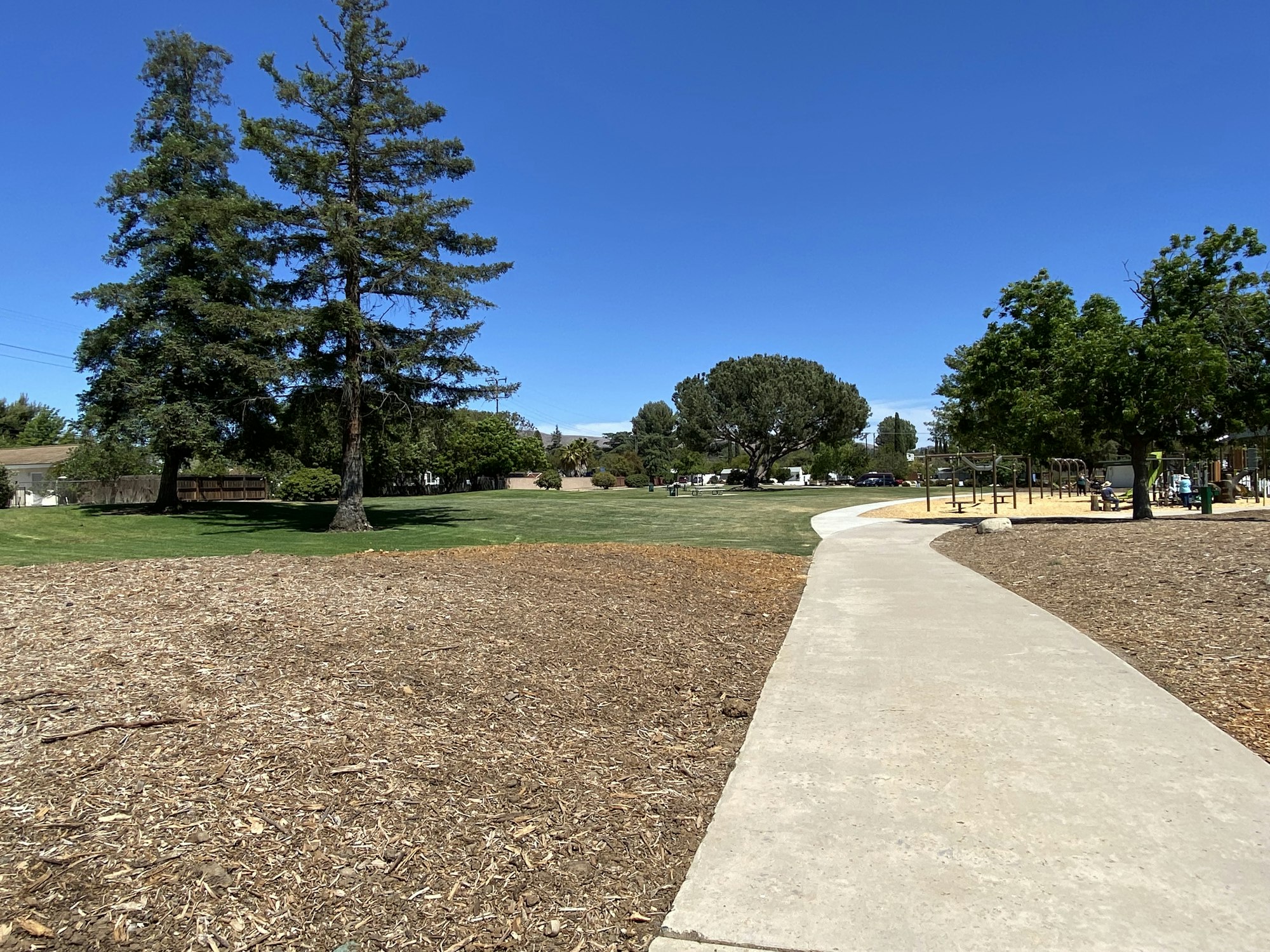 Pathway through a park with trees, a playground, and blue sky.