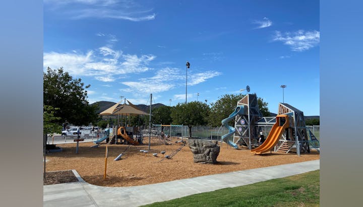 Playground with slides, climbing structures, and shade canopy on wood chip surface under a clear blue sky, surrounded by trees and grass.