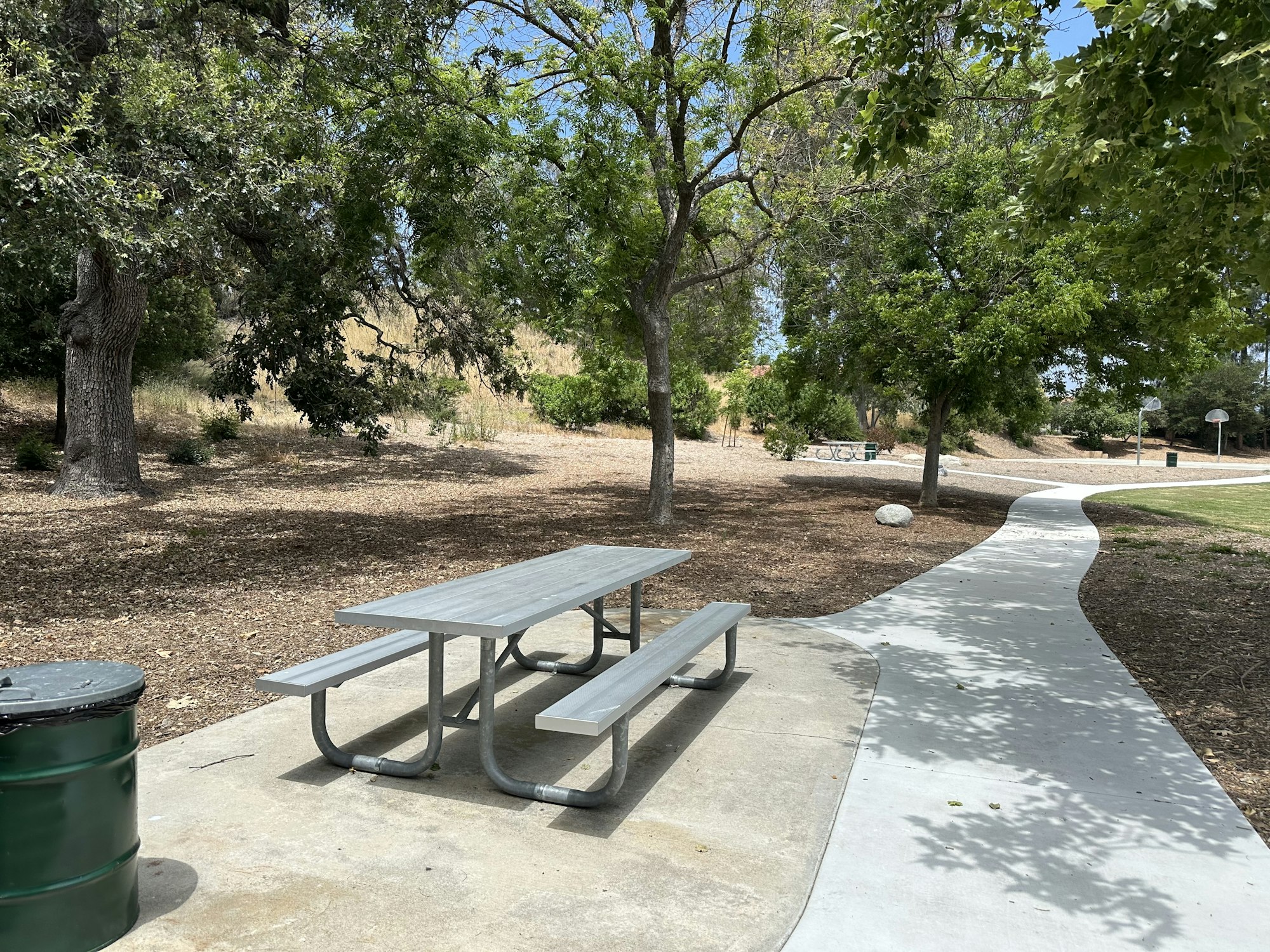 A picnic table under trees in a park, with a walking path and a trash can nearby. Sunny day in a serene environment.