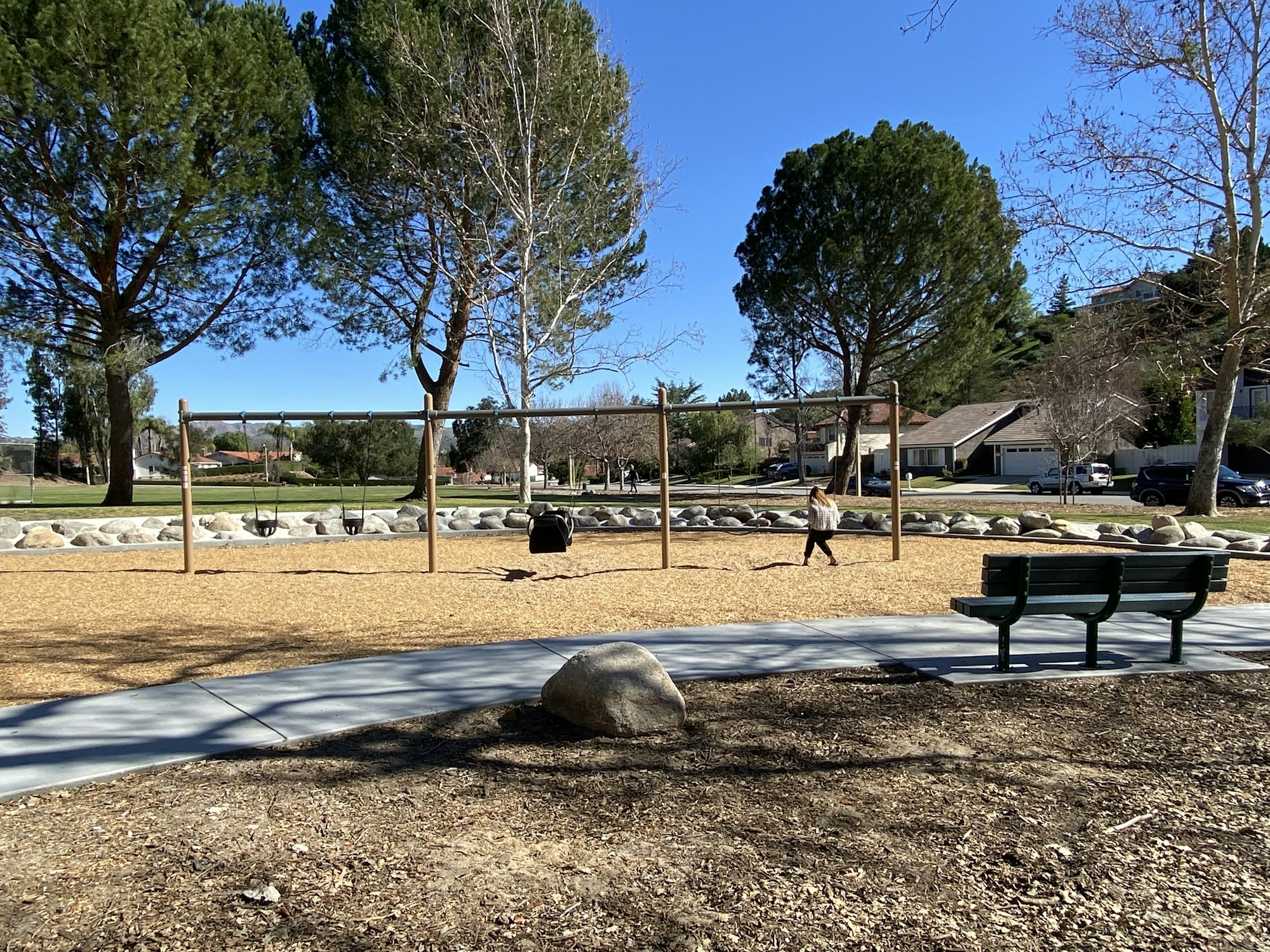 A park with swings, trees, a pathway, and a bench. A person walks near the swing set.