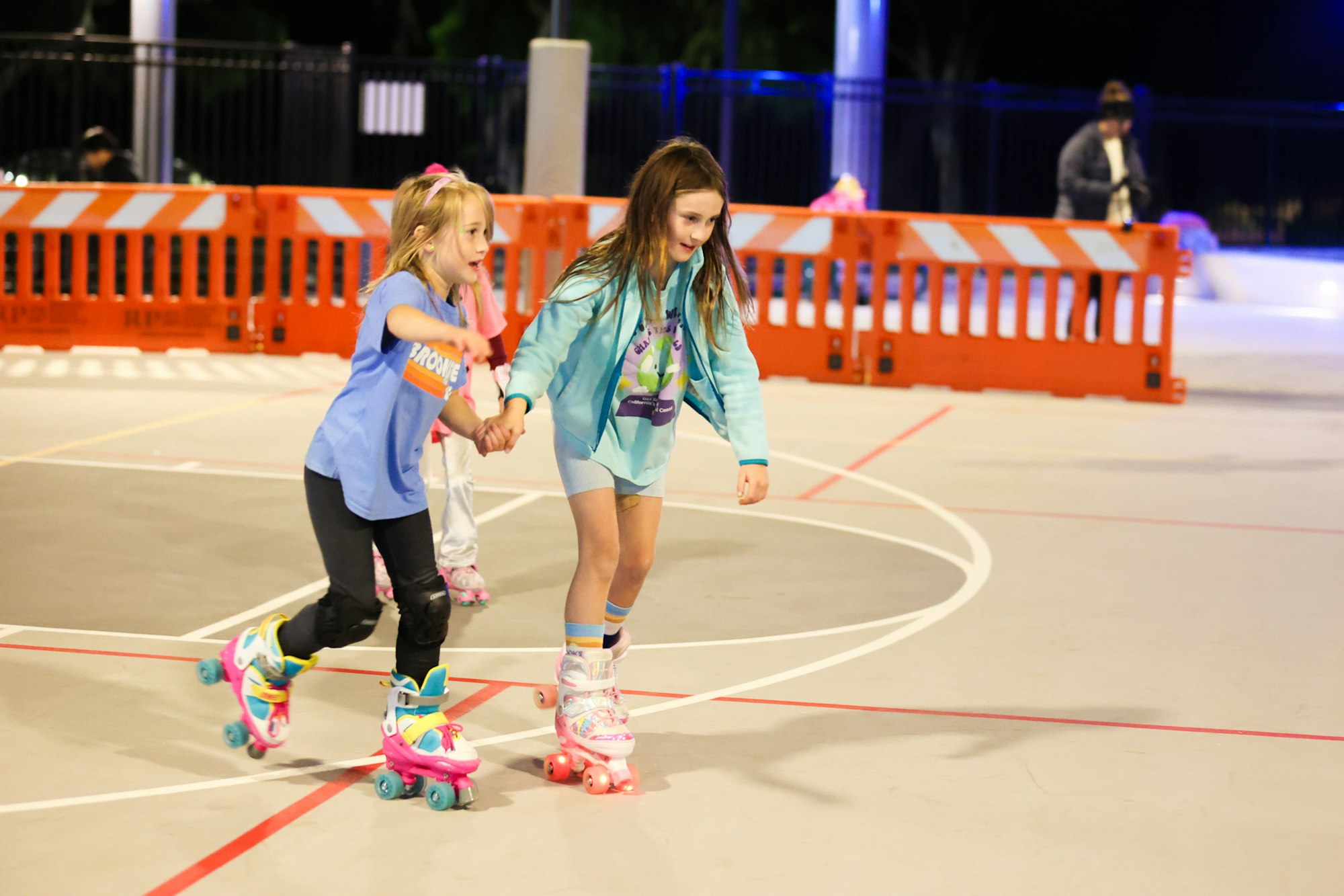 Two girls are roller skating together on a well-lit rink, holding hands and enjoying their time. Safety barriers are visible nearby.