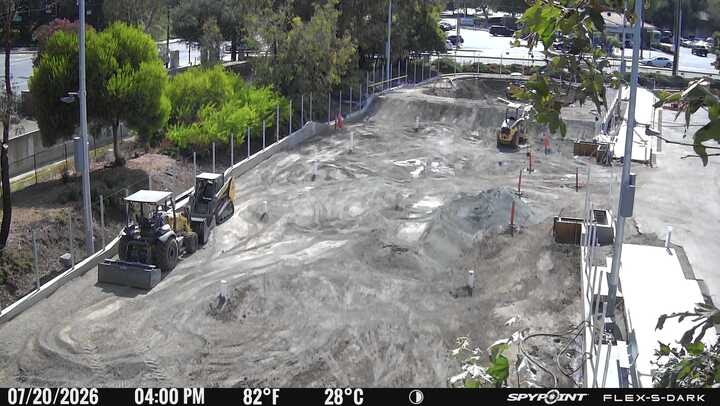 The image shows a construction site with heavy machinery on a dirt surface, surrounded by trees and some infrastructure.