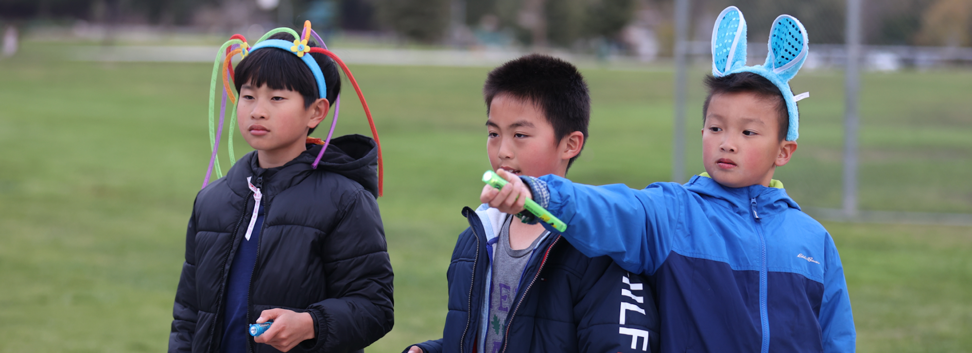 Three kids in jackets outdoors, wearing colorful headbands, one pointing a green tube.
