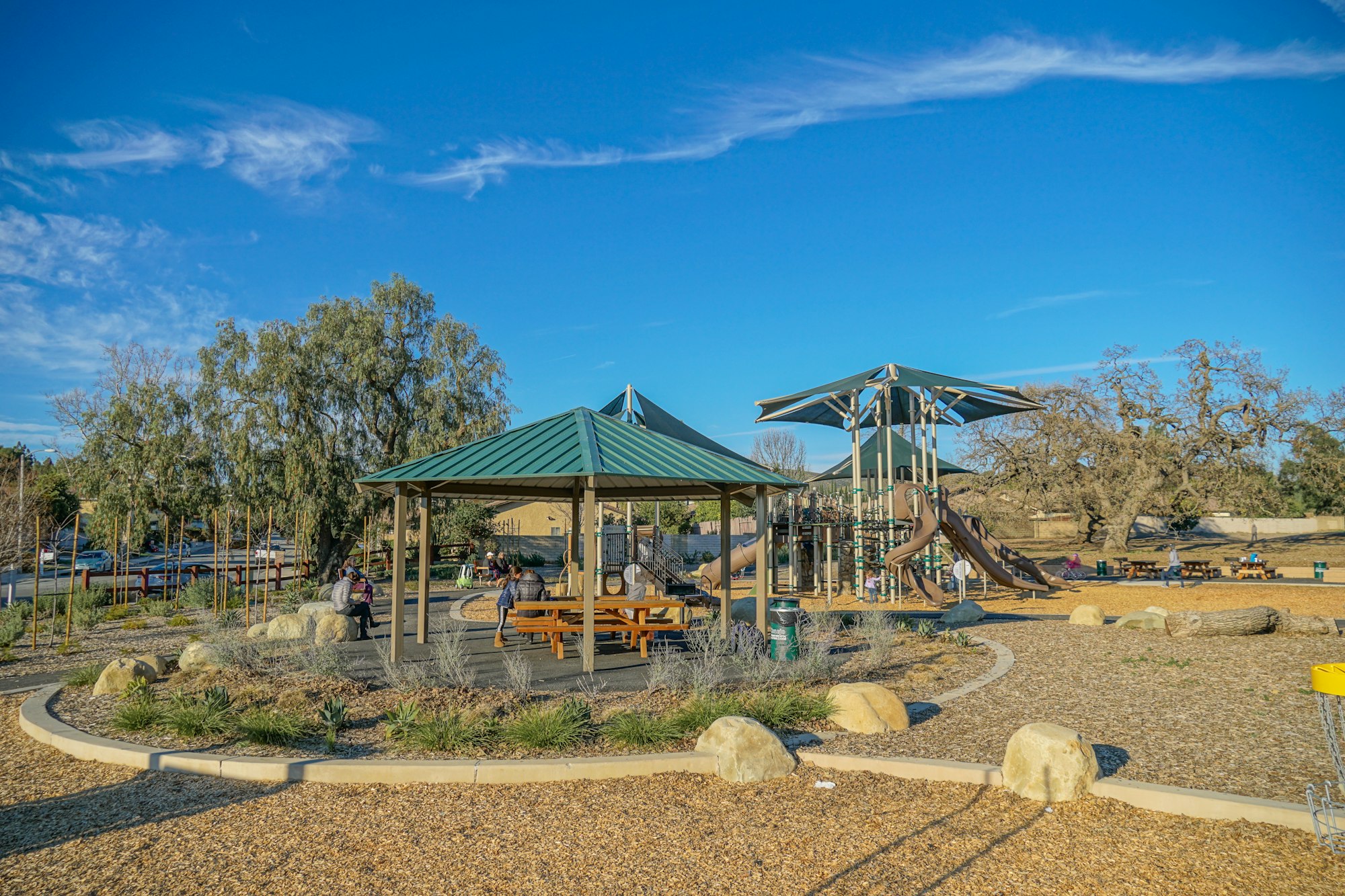 Playground with slides, canopies, a gazebo with seating, trees, and a clear blue sky. Kids and adults are scattered across the area.