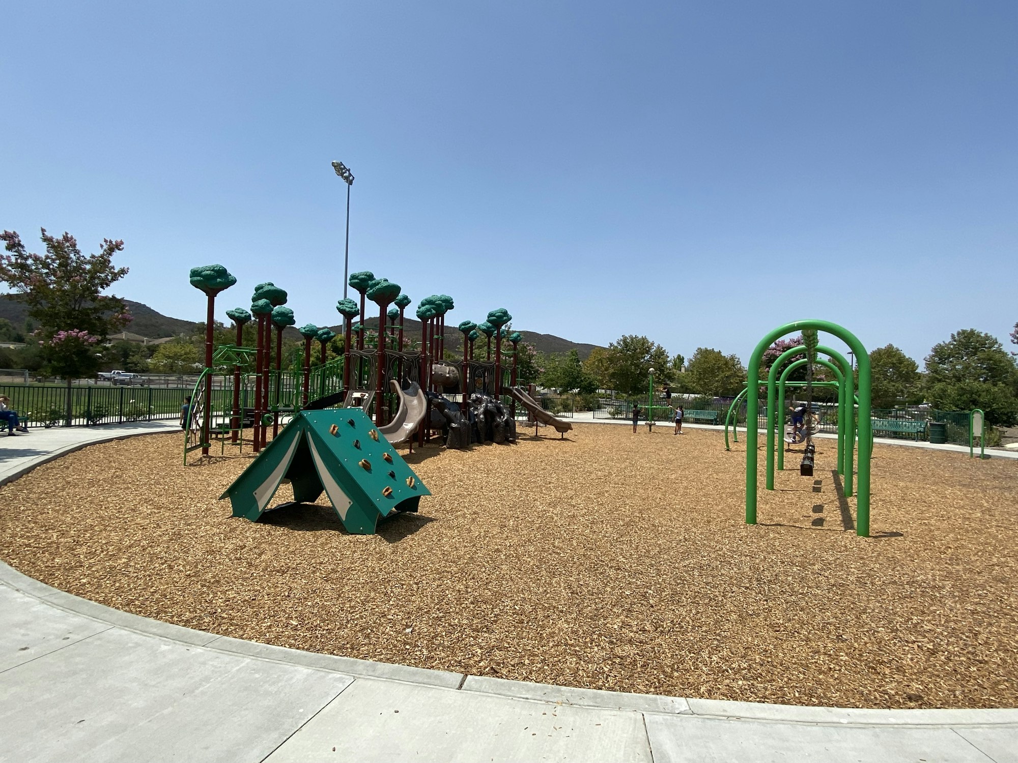 Playground with slides, swings, green structures, wood chips, and trees in the background under a clear sky.