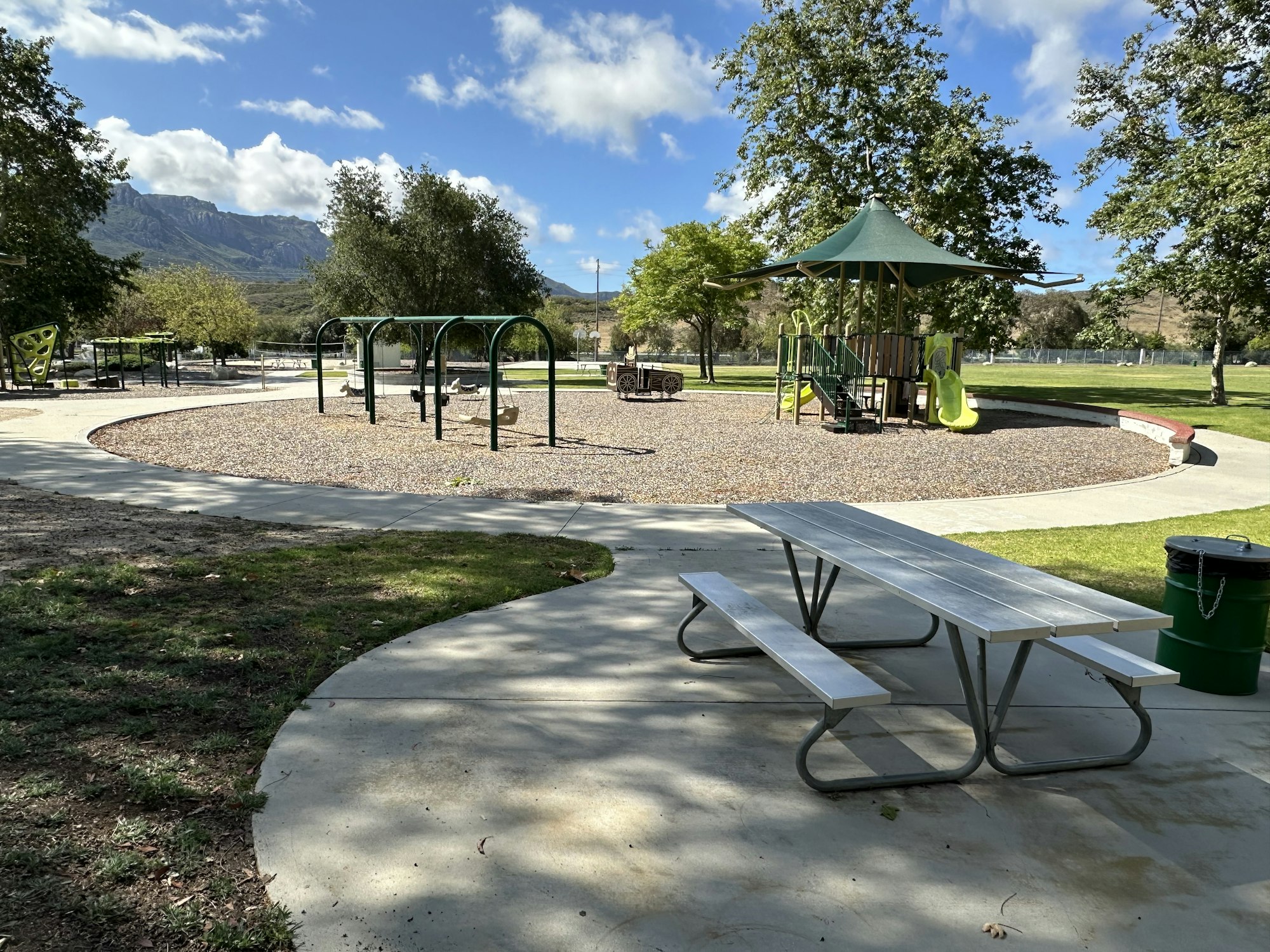 A sunny park scene featuring playground equipment, picnic tables, greenery, and mountains in the background.