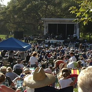 Outdoor concert with a large crowd seated on the grass, facing a stage with performers, surrounded by trees and tents.