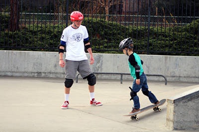 An adult watches a child skateboarding in a park. Both wear helmets and knee pads for safety.