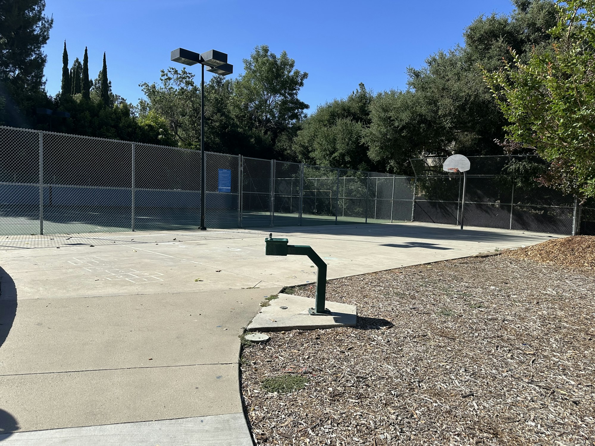 An empty basketball court with a hoop, a watering station, and surrounding greenery under a clear blue sky.