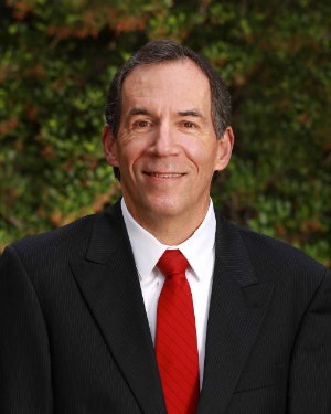 A person in a suit and red tie is smiling with a natural backdrop.