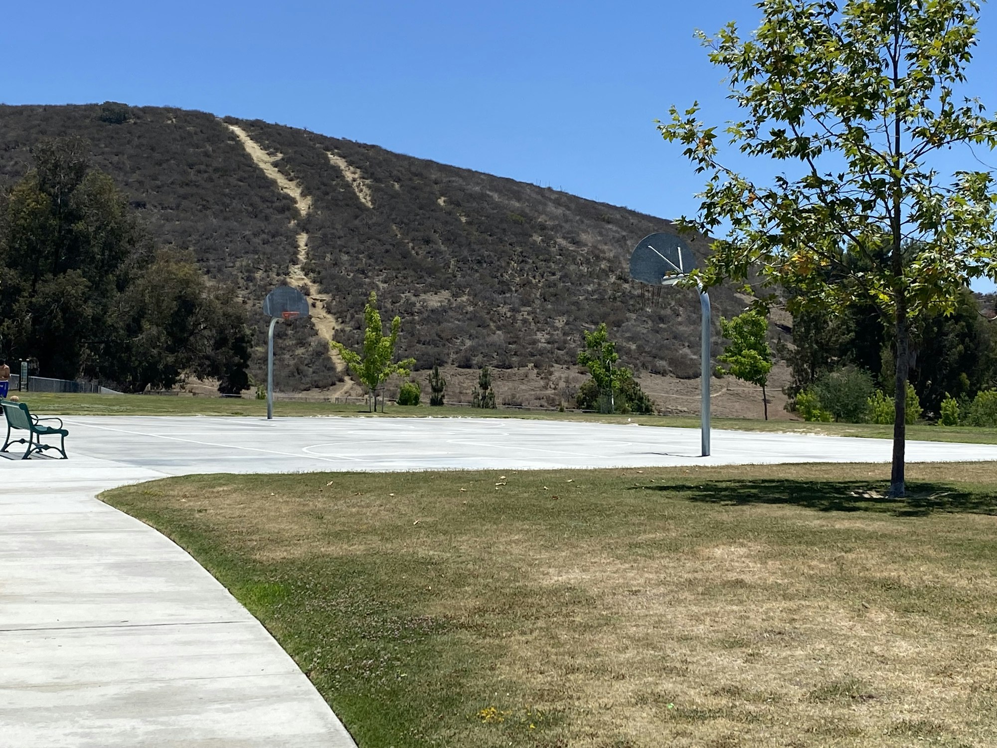Outdoor basketball court with two hoops, nearby benches, a tree, and hills in the background under a clear blue sky.
