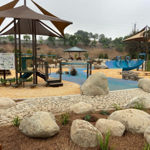 Playground with slides, shade structures, rocks, and a dirt path surrounded by greenery and trees.