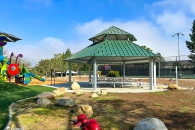A playground with a colorful slide, a pavilion with a green roof, and picnic tables, surrounded by rocks and trees.