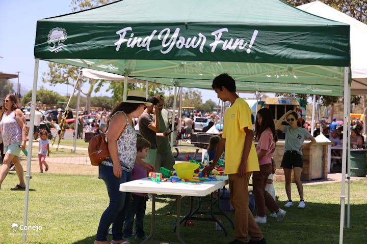People at an outdoor event under a "Find Your Fun!" tent, with tables and toys on the grass.
